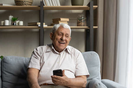 Happy joyful older 80s man holding, using online ecommerce app on mobile phone, resting on sofa at home, chatting on internet, laughing, making call. Distant communication conceptの写真素材