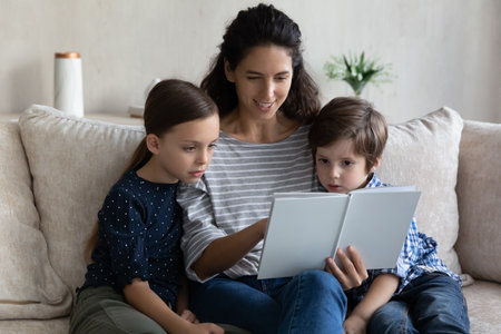 Happy engaged mom reading paper book to son and daughter. Mother and kids sitting on couch at home, enjoying leisure, children listening to exciting fairy tale. Family tome, literature, motherhoodの写真素材