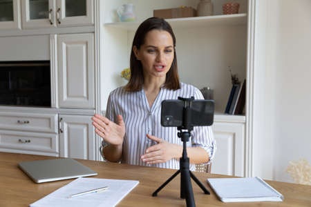 Smiling beautiful millennial 30s woman talking looking at mobile web camera, recording educational video or streaming stories in social network sharing knowledge online, professional blogging concept.の写真素材