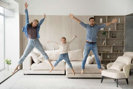 Excited happy active family having fun at home, jumping high in living room, posing for camera in air. Parents and daughter kid dancing to music, singing, screaming, going crazy. Full length portraitの写真素材