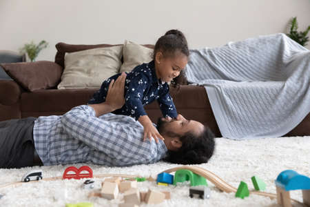 Happy bonding young African American father playing with cute little kid daughter, lying on floor carpet in modern living room. Joyful laughing two generations family having fun on weekend at home.の写真素材
