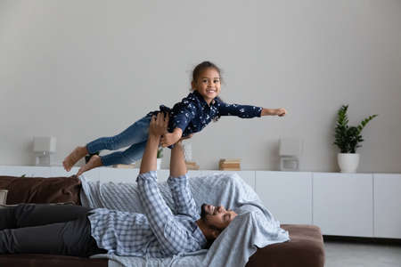 Relaxed millennial happy African American daddy lifting in air laughing cute little kid daughter, imagining travelling or playing plane game on comfortable sofa, enjoying entertaining activity at homeの写真素材
