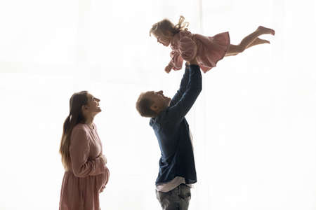 Cheerful expecting parents playing active games with elder child. Dad lifting daughter kid, throwing...