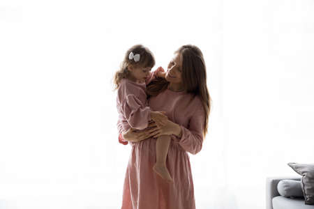 Happy pregnant mom holding elder little daughter girl in arms, smiling, enjoying pregnancy, motherhood. Young expecting mother in pink linen clothes hugging kid, posing against white backgroundの写真素材
