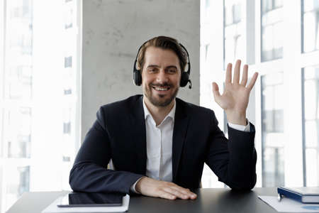 Web camera view smiling pleasant confident young 30s businessman manager worker in formal wear and wireless headset with microphone waving hand, starting distant conversation or recording video.の写真素材