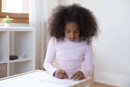 Concentrated cute little teenage curly African American kid girl drawing pictures with colorful pencils on paper alone at home, enjoying creative hobby activity, developing art skills sitting at tableの写真素材