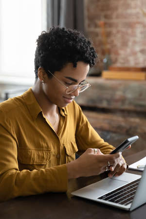 Pensive positive young African gadget addict user woman using cellphone at workplace, sitting at laptop, chatting online, browsing Internet, shopping with ecommerce app. Communication gadgetsの写真素材