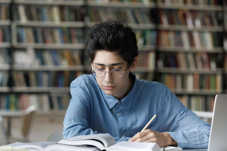 Focused busy young male Jewish student in eyeglasses reading educational textbook, improving knowledge writing notes, preparing for exam or doing homework alone in library, learning concept.の写真素材