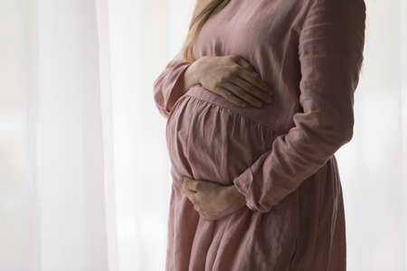 Expecting mom wearing natural dress for pregnant, holding big tummy, feeling kicks, touching and hugging belly with love and care. Childbirth, pregnancy, clothes for expecting concept. Close upの写真素材