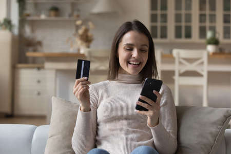 Happy young woman holds smartphone and credit card make purchase remotely, buy goods on internet, transfer money successfully use electronic services. E-shopping, secure pay through e-bank app conceptの写真素材