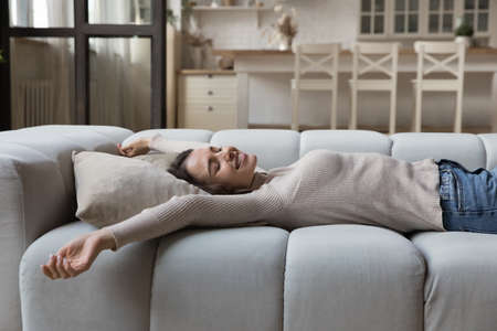 Carefree young woman stretching lying on comfortable couch in living room feeling peaceful, spend...