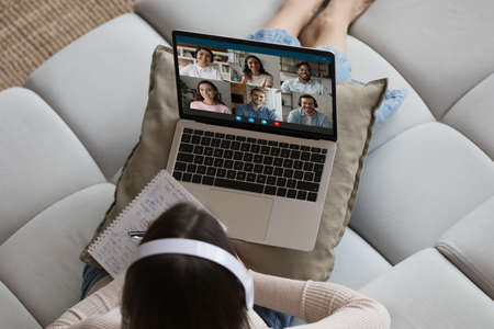 Overhead view woman in headphones sits on sofa takes part in briefing or remote negotiations via video call, multiracial participants, laptop screen view. Virtual meeting event, tech, webinar conceptの写真素材