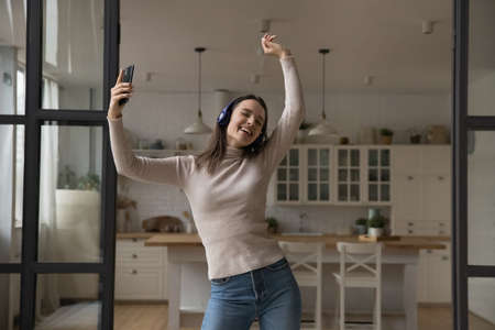 Happy woman wear headphones holds smartphone dance with raised arms, listen song, enjoy favourite track spend carefree leisure at home. Hobby, modern tech, digital music stream service usage conceptの写真素材