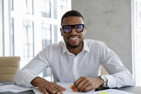 Front view smiling young handsome African American male executive manager leader in eyeglasses wearing wireless earphones holding negotiations video call online meeting with colleagues in office.の写真素材