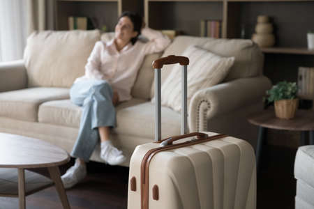 Focus on beige suitcase with blurred carefree young woman sitting on sofa on background. Joyful refreshed millennial Hispanic lady satisfied with vacation travel, relaxing alone in modern hotel room.の写真素材