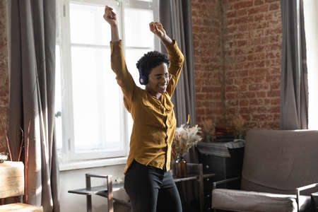 Happy carefree active young African woman in headphones dancing, having fun in loft home interior,...