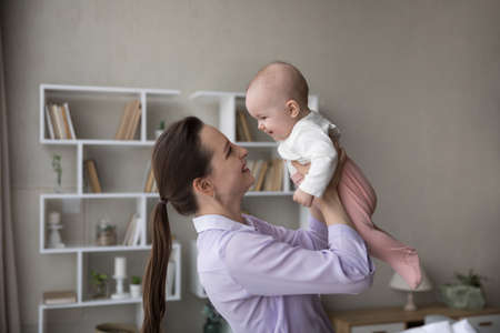 Cheerful happy new mom lifting adorable baby, holding infant in arms, looking at little laughing...
