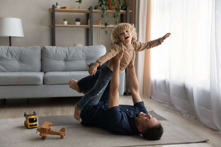 Caring millennial father lying on floor carpet, lifting in air small laughing adorable baby boy, enjoying funny entertaining activity, playing together in living room, daycare, relations concept.の写真素材