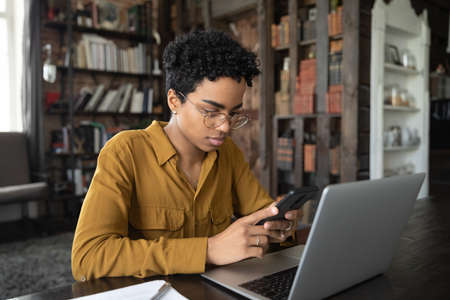 Focused millennial Black freelance employee working at loft home office workplace, using gadgets for online wireless communication, chatting on smartphone at laptop, reading, typing messageの写真素材