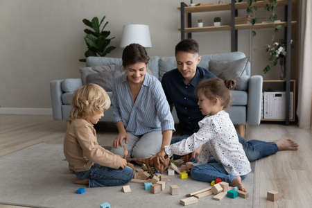Happy bonding caring millennial couple parents and joyful cute kids siblings playing toys, sitting together on carpet on heated warm floor, enjoying entertaining activity in modern living room.の写真素材