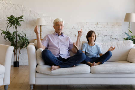 Grandfather and little grandson with eyes closed do meditation practice sit on sofa in living room, make mudra gesture, keep healthy life habit. Lifestyle, hobby of multigenerational relatives conceptの写真素材