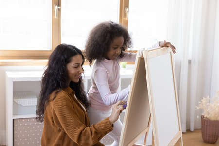 Happy bonding two female generations African American family, young mother and teenage kid daughter drawing with colored felt-tip pens on canvas easel or white board, enjoying hobby activity concept.の写真素材