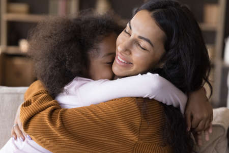 Happy loving little preteen curly child girl cuddling smiling African American young mother, missing after long separation time, showing affectionate feelings, enjoying sweet tender moment at home.の写真素材