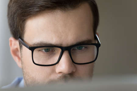 Close up face concentrated millennial male scientist engineer businessman in eyeglasses look on computer screen do research job. Serious young executive work by pc think on business project read emailの写真素材