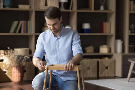 Skilled millennial guy engaged in do it yourself activity construct furniture from wooden details. Busy young man husband collect shelf table from parts furnish decorate home with self assembled craftの写真素材