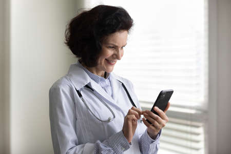 Positive satisfied mature head doctor woman using smartphone in office, chatting with patients with medical app, giving online consultation, texting message on cellphone, reading healthcare newsの写真素材