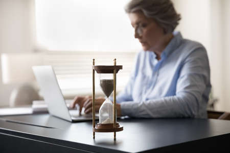 Busy productive mature businesswoman office employee work on laptop sit at workplace desk, hourglass...