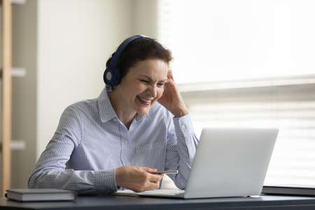 Happy senior student woman in headphones watching webinar on laptop, talking to teacher, coach in...