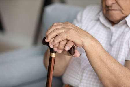 Close up cropped view disabled older man holds cane looks desperate or lonely sit on sofa in nursing...