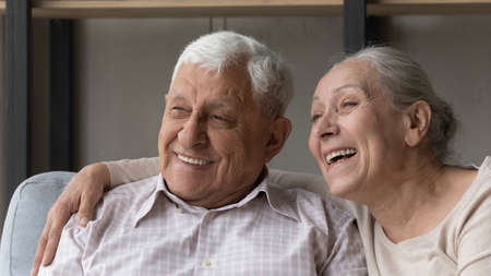 Close up portrait grey-haired spouses sit on sofa smile staring into distance look happy. Marriage, harmony and love between senior wife and husband, medical insurance cover for older citizen conceptの写真素材