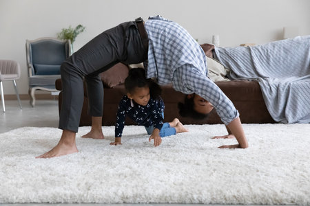 Active African yogi dad hold bridge stand on white soft carpet, doing yoga, gymnastics, exercising at home, watching little daughter girl crawling around. Father and kid playing active gamesの写真素材