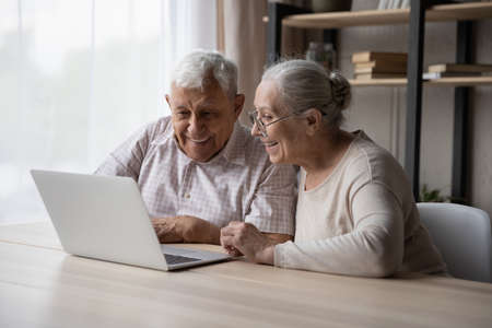 Happy older couple sit at table at home staring at laptop screen enjoy easy comfort usage of modern tech, make online purchase, buy goods, electronic services through e-commerce, technologies conceptの写真素材