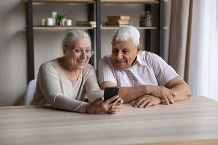 Smiling older couple sit at table staring at smart phone screen, enjoy new mobile application, lead online chat in social media with grown up children, make order, buying through e-commerce conceptの写真素材