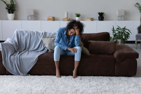 Depressed exhausted young African woman feeling stress, suffering from headache, sitting on cough at...