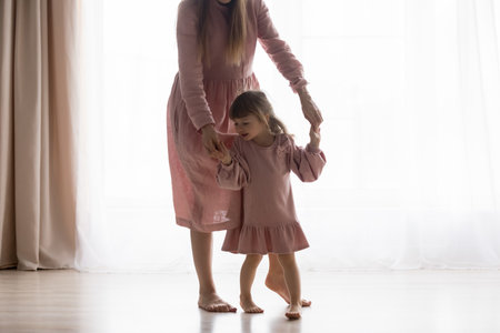 Caring young mother holding hands of small kid daughter, helping making steps barefoot on warm heated wooden floor, helping kid daughter with walking disability or injury, rehabilitation concept.の写真素材