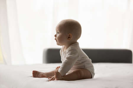 Side view happy cute sweet small newborn baby boy girl sitting on bed, looking away. Cheerful...