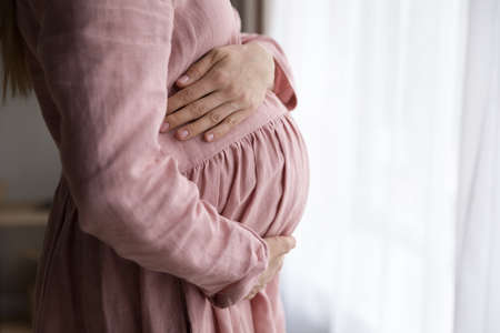 Cropped side view dreamy young pregnant woman standing near window, feeling inspired waiting for first baby, stroking big belly, thinking of future labor, daydreaming at home, IVF advertisement.の写真素材