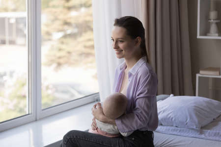 Smiling dreamy young mother looking in distance while breastfeeding little newborn baby boy girl,...