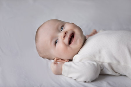 Head shot close up happy small cute kid boy girl in white bodysuit lying on bed, laughing looking away, having fun after waking up, entertaining alone in bedroom, carefree childhood, baby care conceptの写真素材