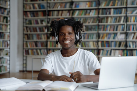 Portrait of African guy student enjoy e-learns use modern tech, sit at table in library wear headphones look at camera. Education, studies, gain new skills, improve foreign language knowledge conceptの写真素材