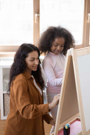 Positive focused daycare teacher helping little student kid, school pupil, kindergarten African child to draw at small easel, childish whiteboard. Mom and daughter enjoying education activity at homeの写真素材