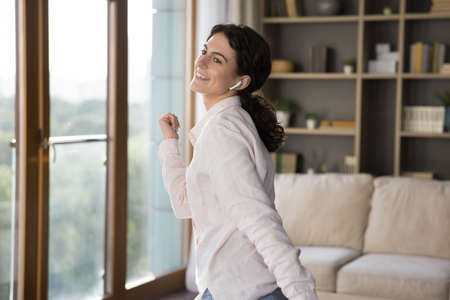 Happy joyful active Latin girl with wireless earphone in ear enjoying favorite songs, tunes, dancing in living room, listening to music with online app, service, smiling. Cheerful woman at home partyの写真素材
