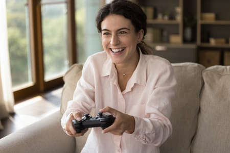 Happy cheerful young gamer girl reducing stress, enjoying exciting videogame, playing video game, holding gamepad, using joystick, game controller, laughing, sitting on home sofaの写真素材