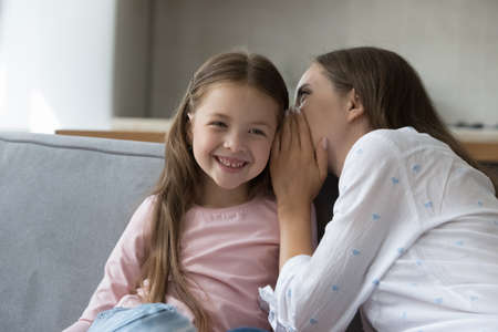 Happy mom whispering in girls ear, sharing secret with joyful kid. Young mother telling funny story...