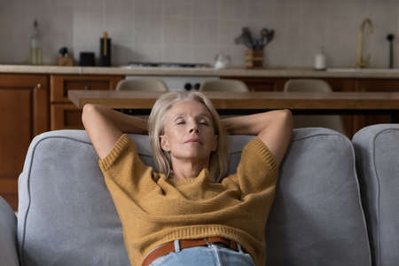 Calm peaceful elder lady relaxing with closed eyes on comfortable sofa at home, taking deep breath...