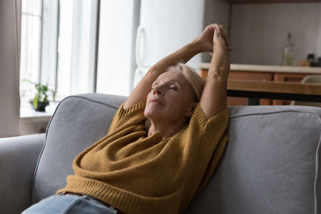 Peaceful relaxed senior lady stretching body on comfortable sofa, breathing fresh cool purified air,...